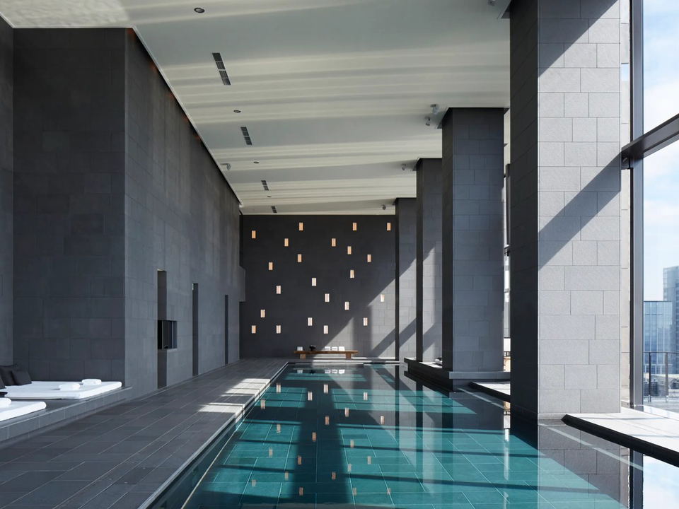 A serene, minimalist indoor lap pool at Aman Tokyo's spa, featuring dark stone walls, a teal-tiled pool floor, and white lounge chairs.