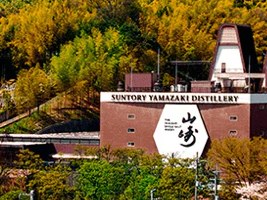 A wide landscape view of the Suntory Yamazaki Distillery nestled among lush green and golden forested hills, with a tall transmission tower visible.