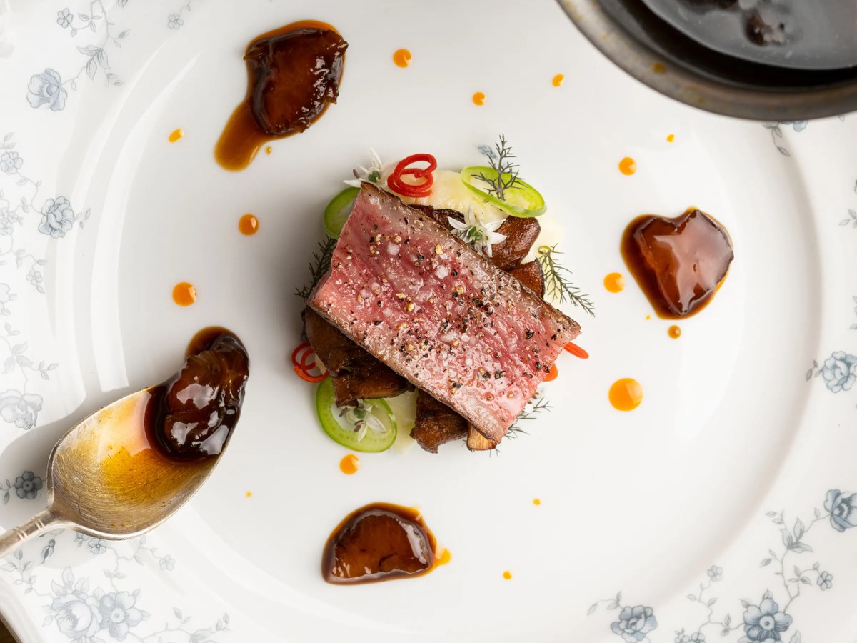 A beautifully plated dish of A5 strip loin with potatoes, black garlic, and braised tendon on a white plate with blue floral patterns.