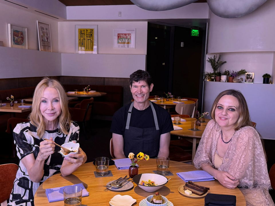 A chef in a black apron stands between two seated women at a restaurant table with plates of food, artwork on walls, and an exit sign visible in the background.