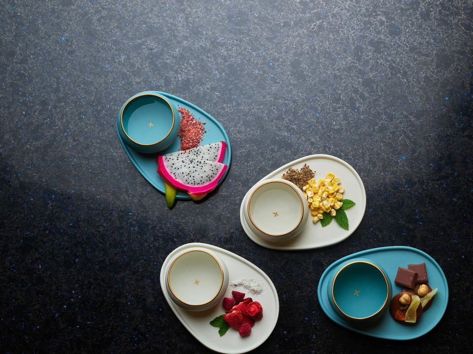 Four oval trays, two teal and two cream, each with a small bowl and various food items like dragon fruit, corn, raspberries, and chocolate.
