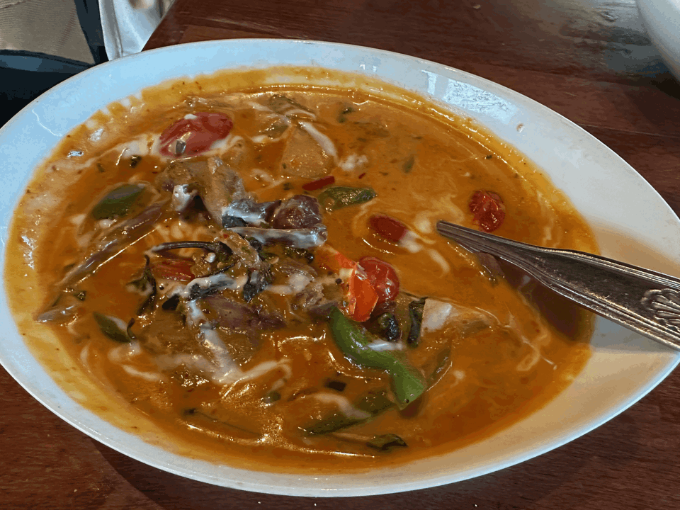 A close-up shot of a white bowl filled with a vibrant orange curry, featuring chunks of meat, green vegetables, and halved cherry tomatoes.