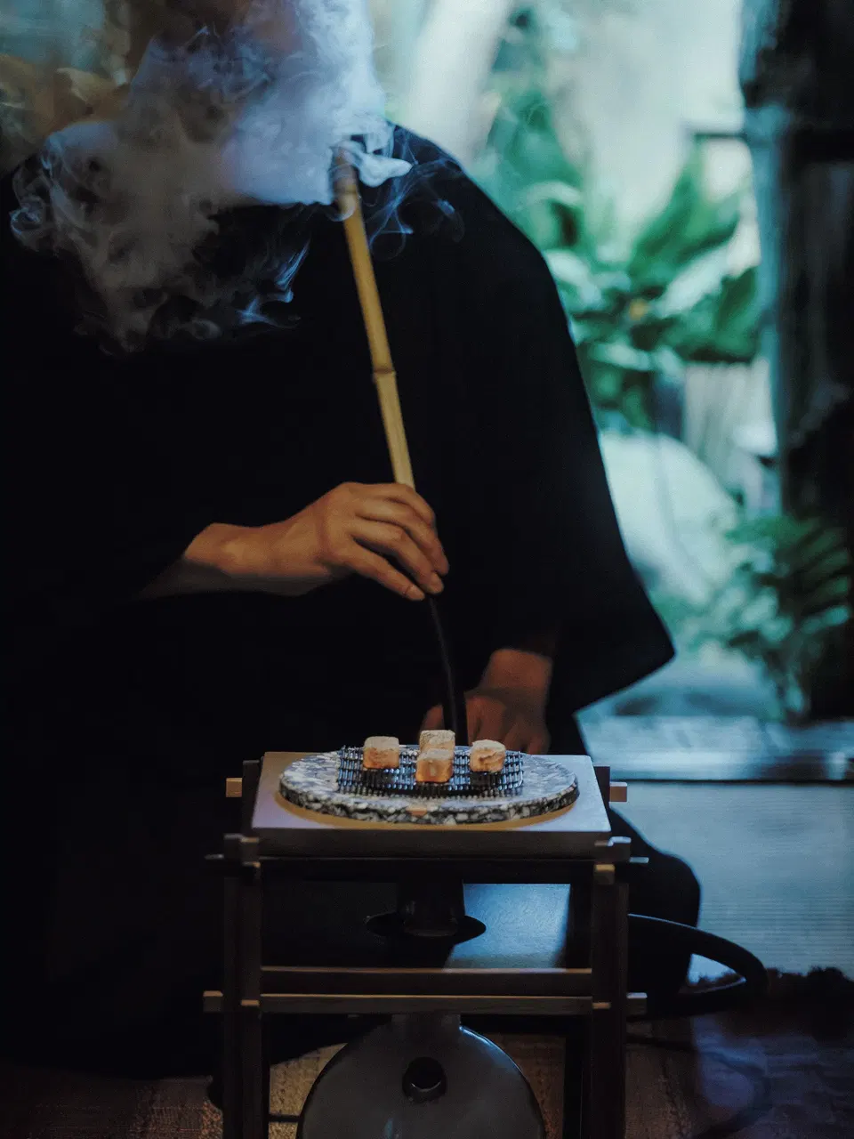 A person in dark clothing holds a bamboo whisk over a ceramic tea bowl on a black stand, with incense smoke visible and blurred green elements in the background.