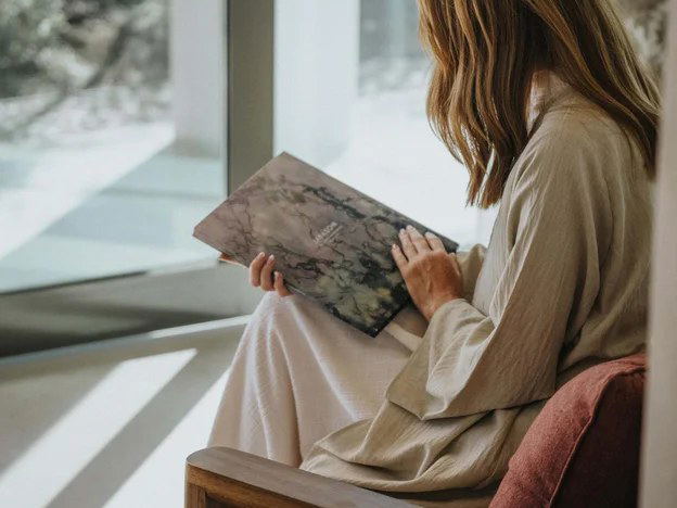 A woman in neutral-toned clothing sits in a modern, light-filled interior space holding what appears to be a wellness journal or consultation material