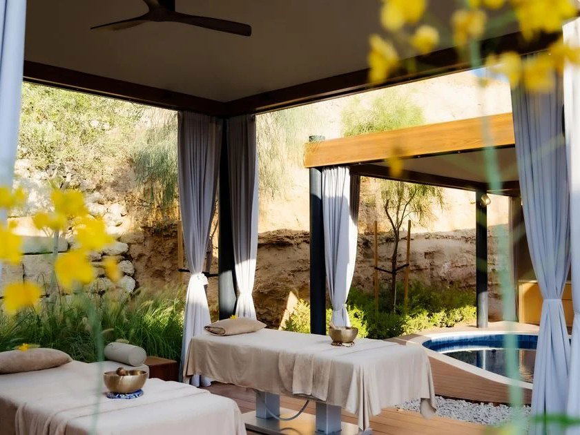 Outdoor spa treatment pavilions with massage tables, white curtains, and a pool, set in a landscaped courtyard with modern architecture; source page e