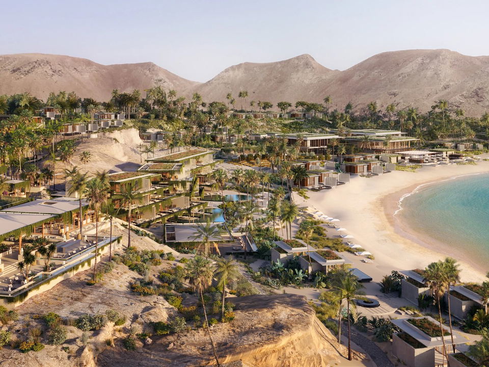 Aerial view of a luxury beachfront resort development with modern villas, palm trees, sandy beach, turquoise water, and arid mountains in the backgrou