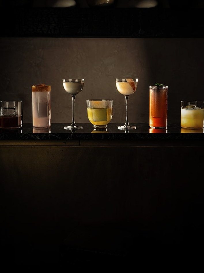 Seven different cocktails in various glass types arranged in a line on a black bar surface against a dark background, displaying amber, golden, and clear colored beverages.