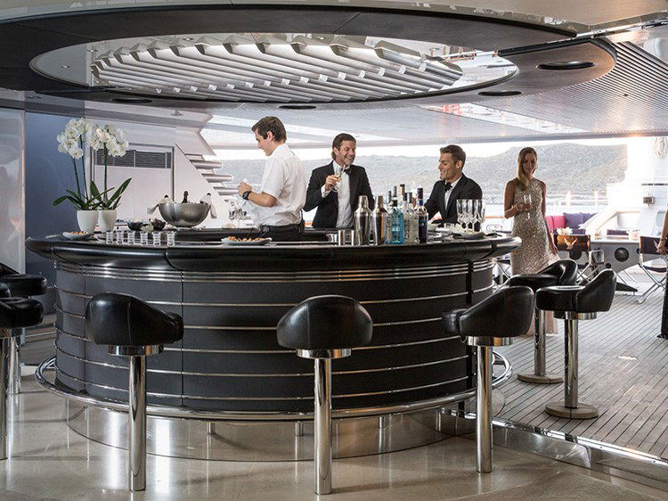 A circular black bar with chrome accents and black leather stools on the deck of the Maltese Falcon superyacht, with guests and a bartender.