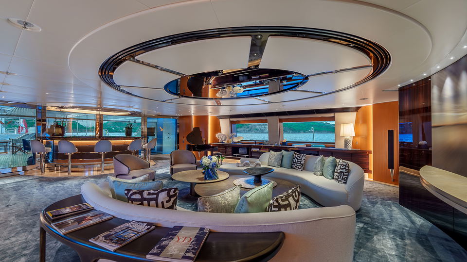 Main salon interior of the Maltese Falcon superyacht, showing curved cream sofas, patterned cushions, a grand piano, and a large circular mirrored ceiling feature with harbour views through the panoramic windows.