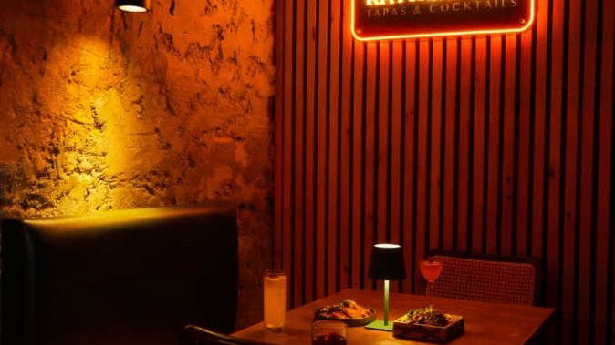 A warmly lit, intimate booth interior at Kathmandu Tapas & Cocktails, featuring a glowing neon sign and a table with drinks and food.