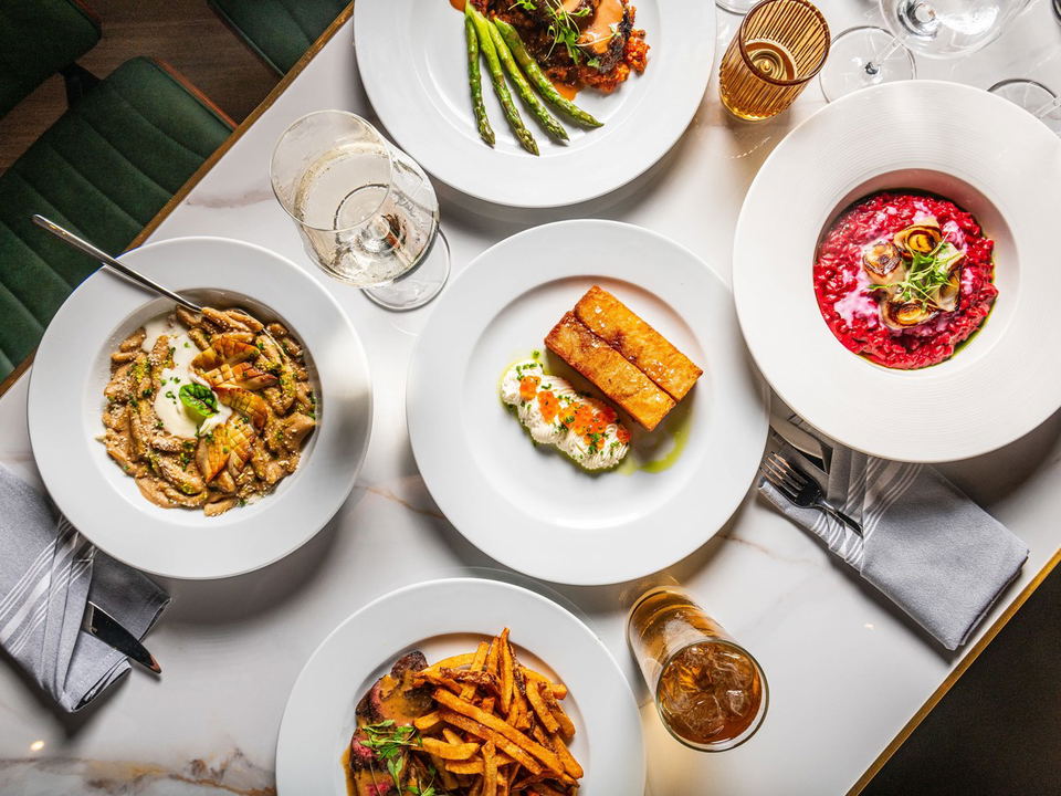 Overhead flat-lay of five plated dishes on a white marble table including king mushroom cavatelli, red beet risotto, french fries with steak, crispy