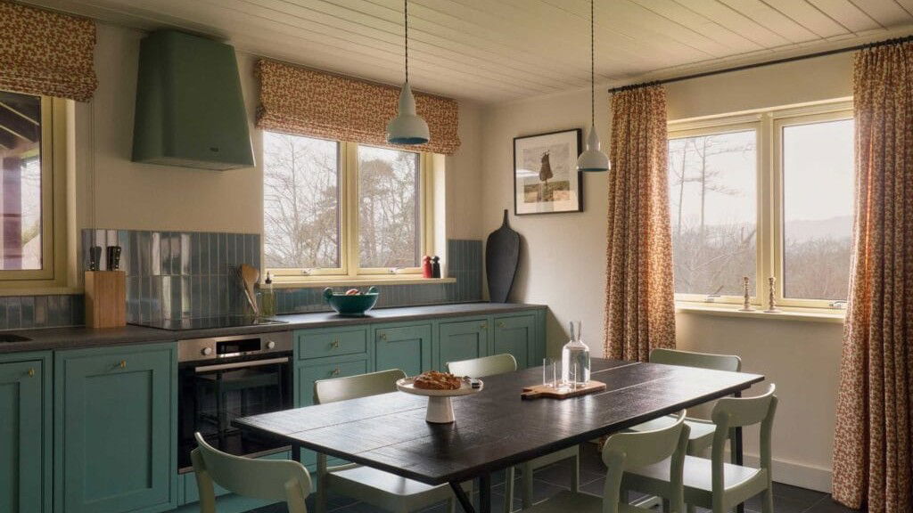 A modern kitchen and dining room interior with sage-green cabinetry, dark wood dining table, pendant lighting, and patterned curtains overlooking a wi