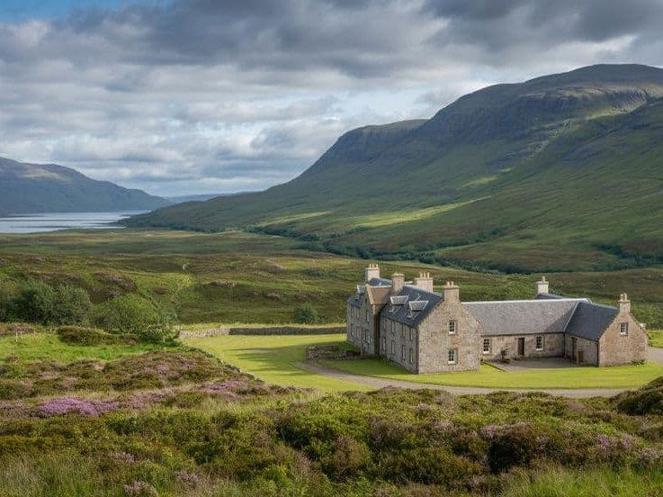 Wide exterior shot of restored stone cottages with multiple chimneys set against dramatic Scottish Highland landscape with mountains, water, and moorl