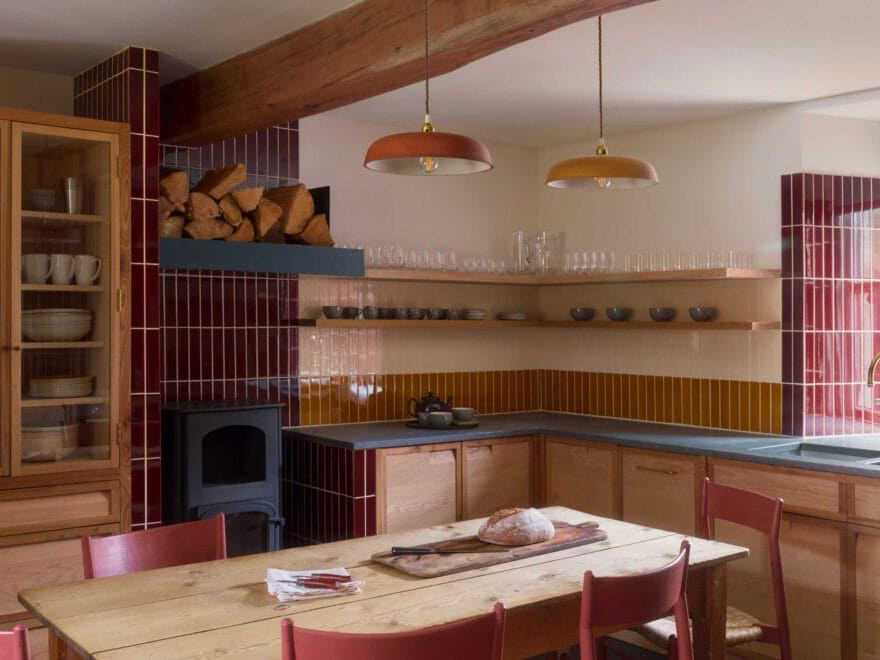 A kitchen interior with light wood cabinets, burgundy and mustard vertical tiles, open shelving, copper pendant lights, and a dining table with pink chairs.