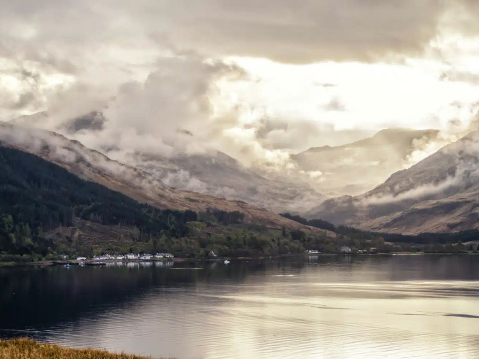 A scenic view of a loch with mountains in the background under cloudy skies, with a small village visible along the far shore.
