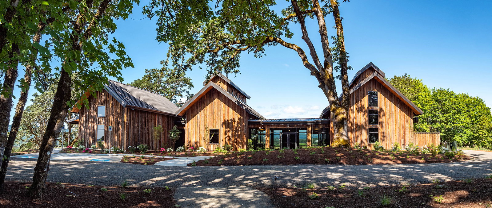 Three wooden buildings with dark roofs and large windows are nestled among trees under a clear blue sky, connected by a glass-enclosed walkway.