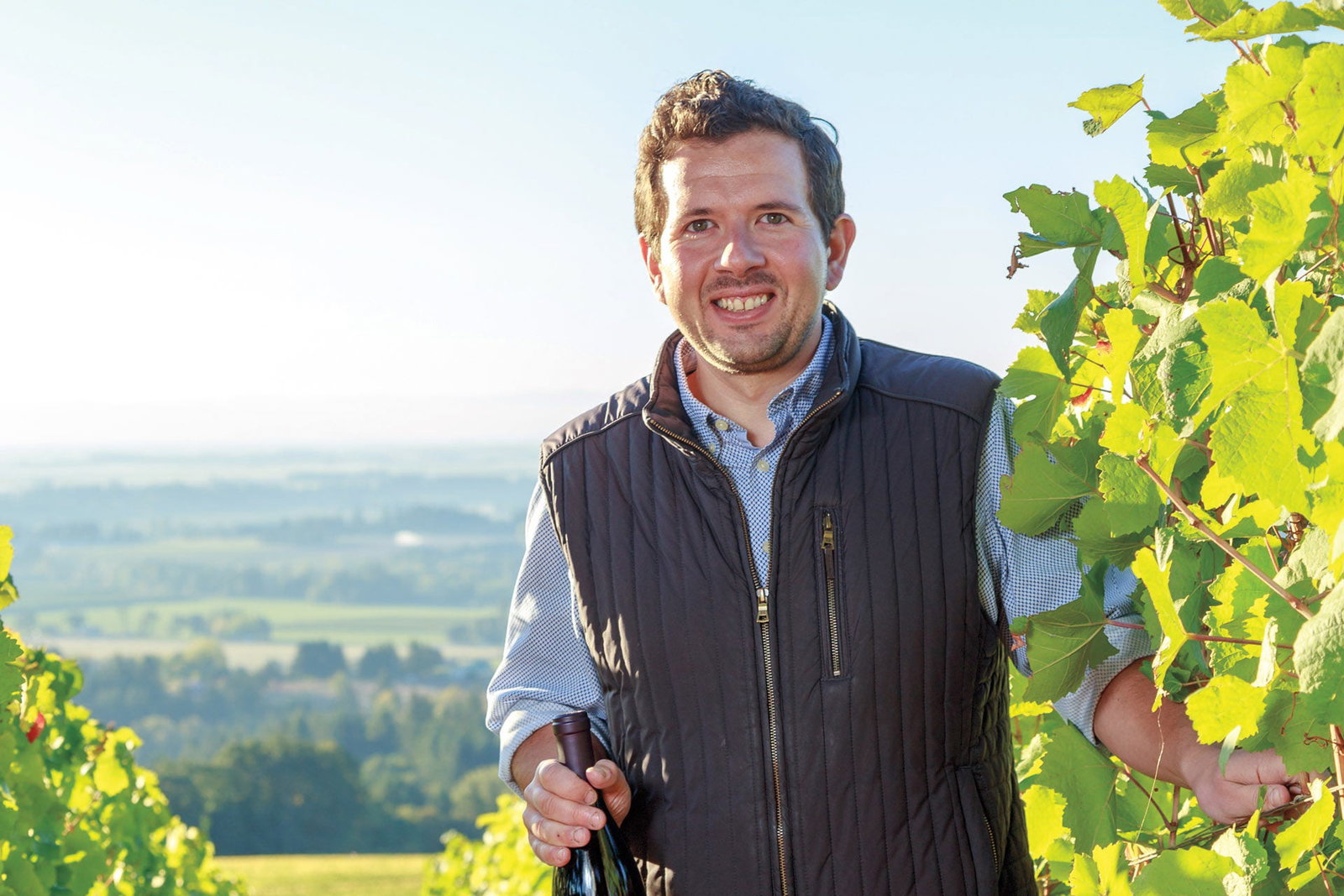 Smiling man in a quilted vest holding a wine bottle among grapevines at Koosah Vineyard, Oregon, with a valley view behind him.