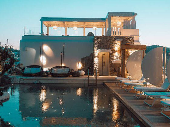 A dusk exterior shot of the Kove Hotel Mykonos MGallery, featuring a pool with reflections, sun loungers, and an illuminated upper terrace.