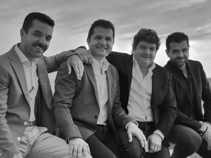 Black-and-white photo of four smiling men in blazers seated outdoors, left to right Panos, Markos, Vangelis, and Marios Daktylides, with a coastal sky behind them.