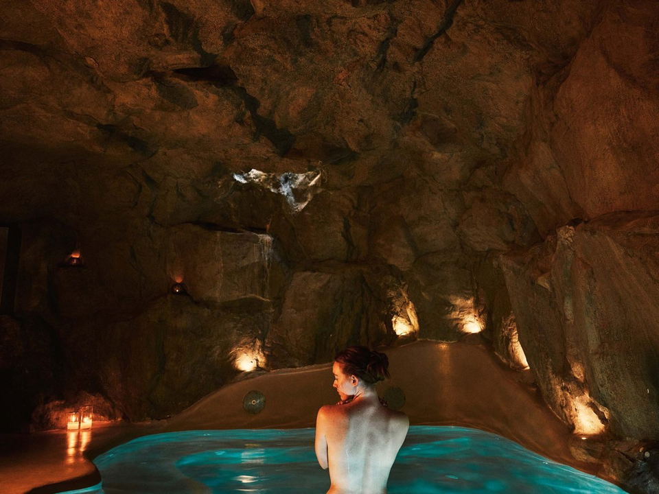 A woman with her back to the camera, shoulders bare, soaking in a glowing turquoise pool inside a dramatic natural rock cave environment with warm