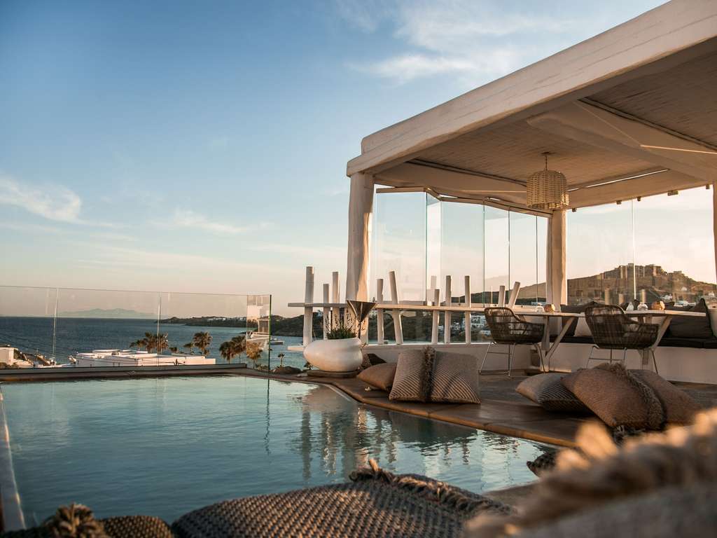 An infinity pool overlooks the sea and a distant town, with a covered outdoor dining area featuring wicker chairs and cushions.