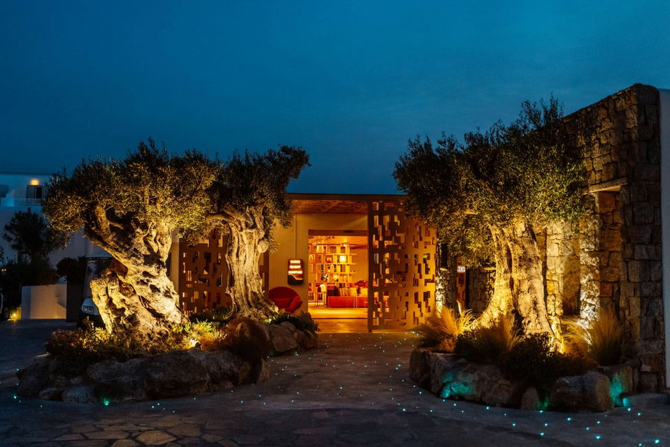 Exterior entrance of Kove Hotel Mykonos MGallery at dusk, with two ancient uplighted olive trees flanking corten steel gates, turquoise fiber-optic lights in the stone forecourt, and a warmly lit lobby visible beyond.