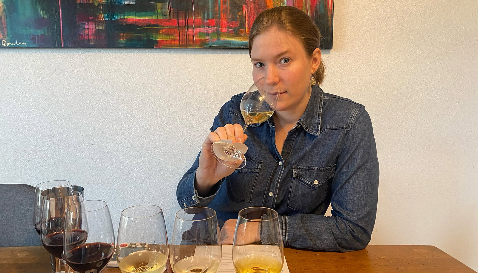 A woman in a denim shirt conducting a blind wine tasting with multiple numbered glasses arranged on a table.