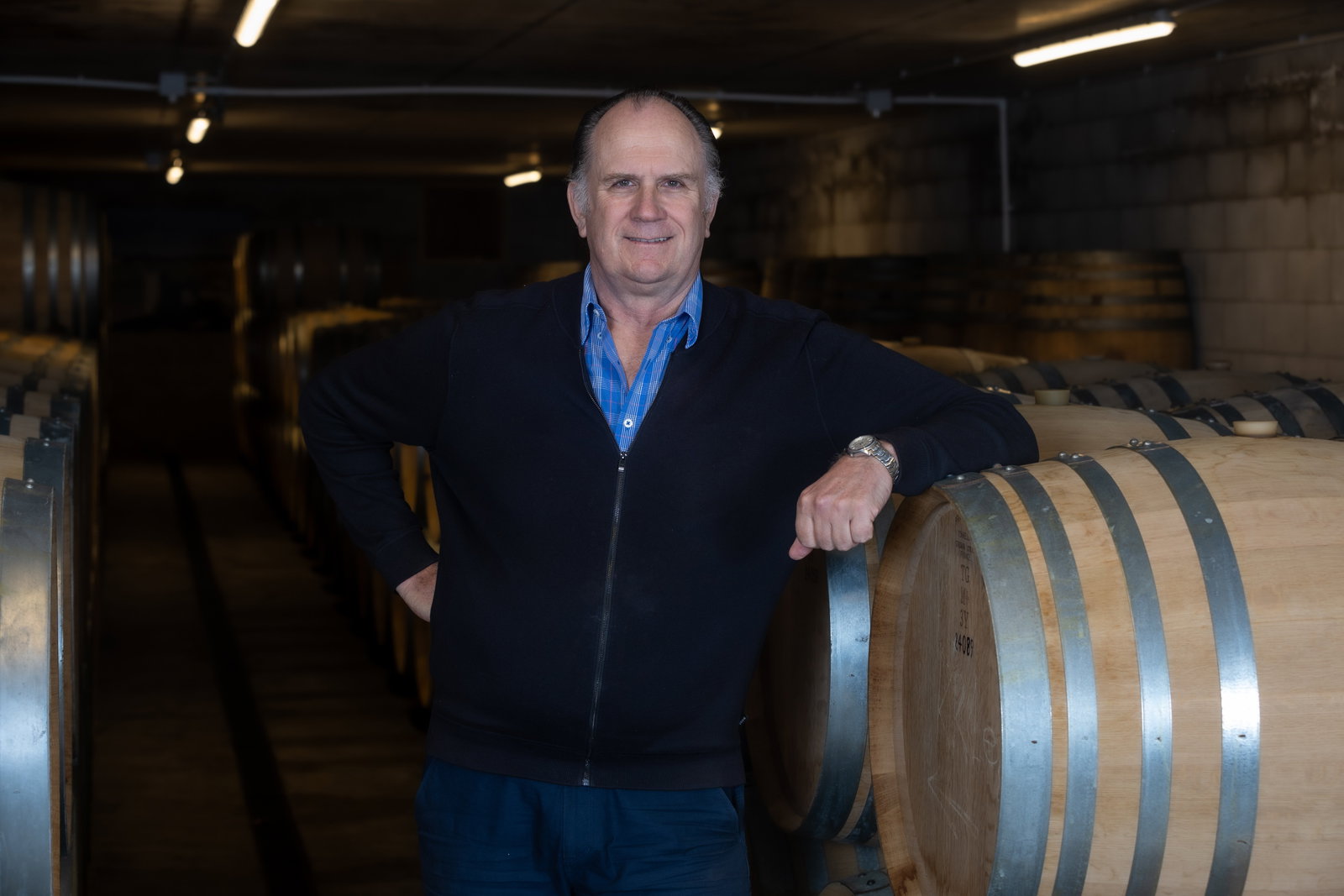 A man in business casual attire (blue shirt, dark zip-up sweater) standing in a wine cellar with wooden barrels visible in the background.