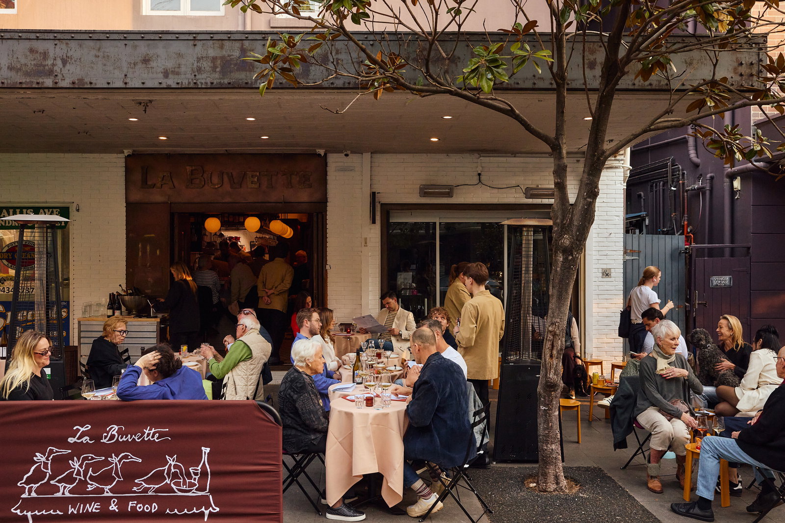 Outdoor restaurant scene at La Buvette with diners seated at white-clothed tables, staff moving between indoor and outdoor spaces, and the restaurant's signage visible on the storefront.