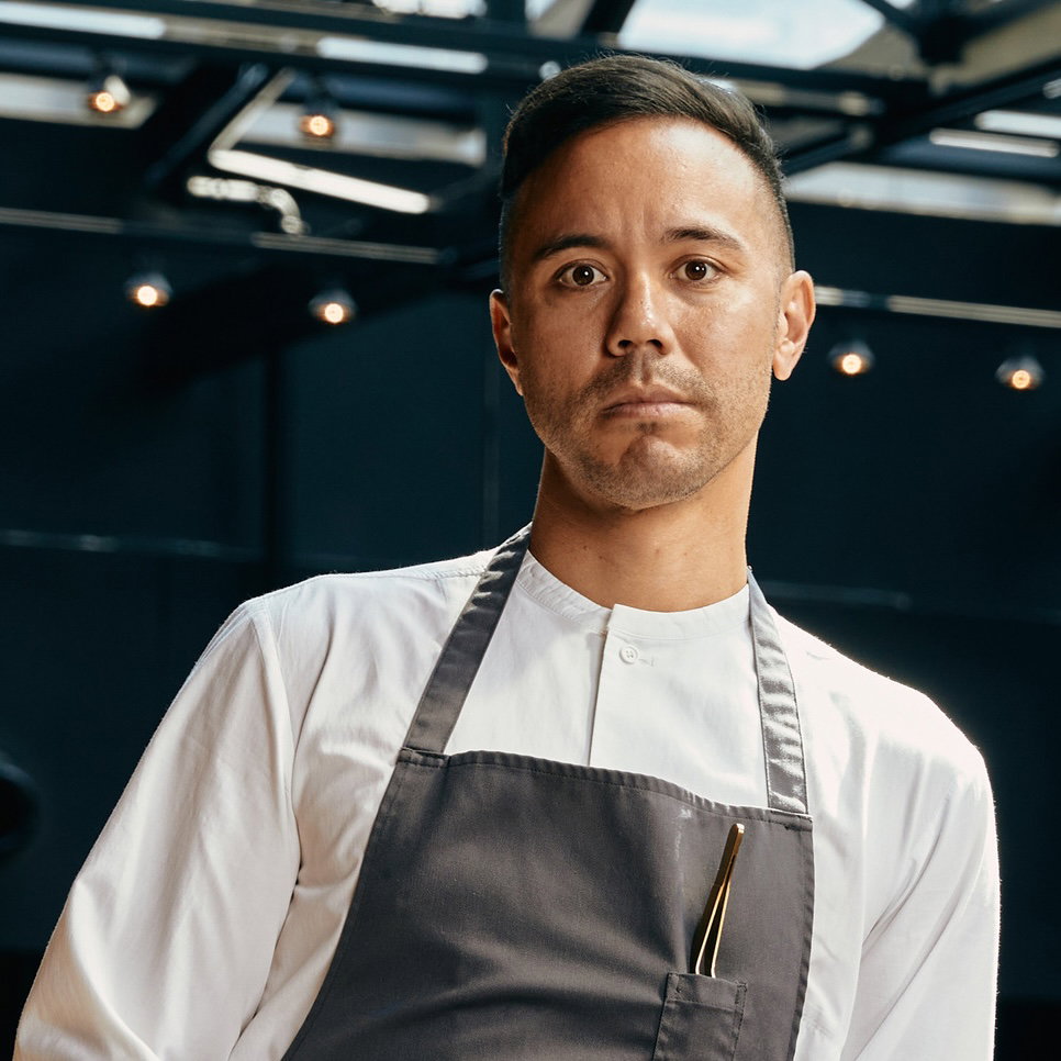Professional portrait of a chef in white chef's coat and gray apron with pen in pocket, photographed in a modern restaurant setting; source page expli