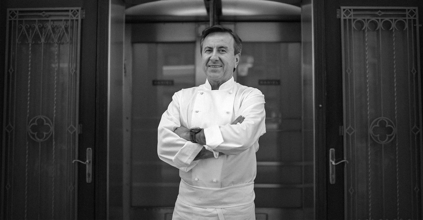 Professional portrait of a chef in white chef's coat standing with arms crossed in front of ornate doors