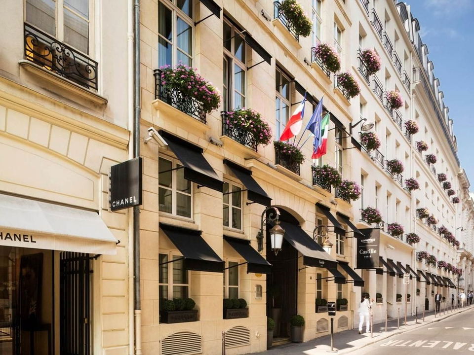 The exterior of Hotel Castille Paris, featuring black awnings, flower-filled balconies, and a 'Castille Paris' sign, adjacent to a 'CHANEL' boutique.