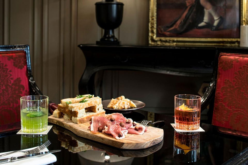 Two drinks, one green and one amber, on a black table with sandwiches and charcuterie. The green drink has a mint leaf, and the amber drink has an