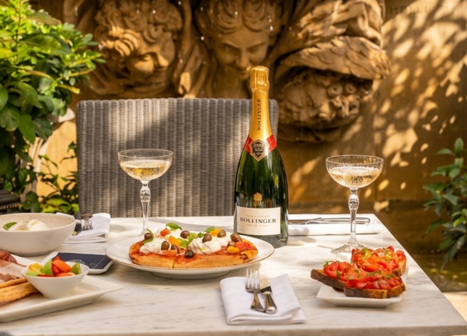 A bottle of Bollinger champagne and two glasses of champagne on a marble table with various food items, including a flatbread pizza and bruschetta,