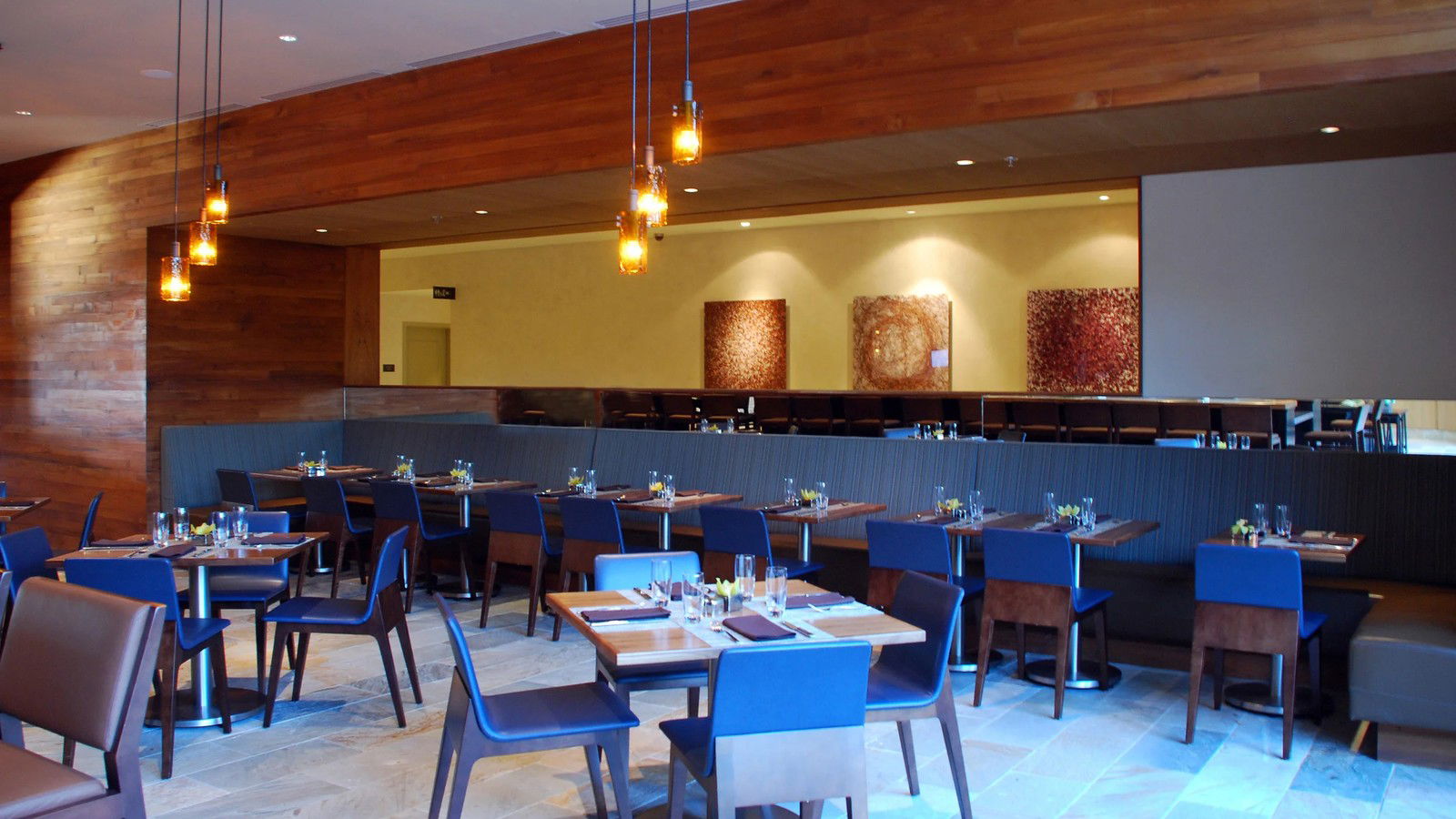 A modern dining room with blue chairs, wooden tables set with glassware, pendant lights, and abstract artwork.