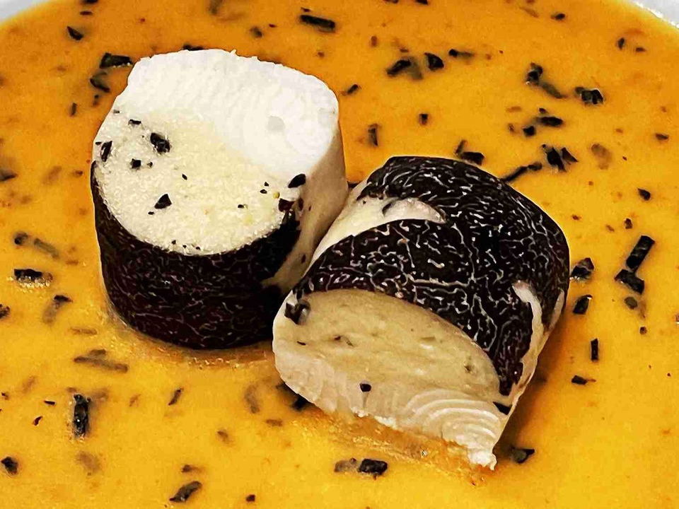 Two cylindrical rounds of white cheese wrapped in black truffle slices, served in a golden bisque with minced black truffle.