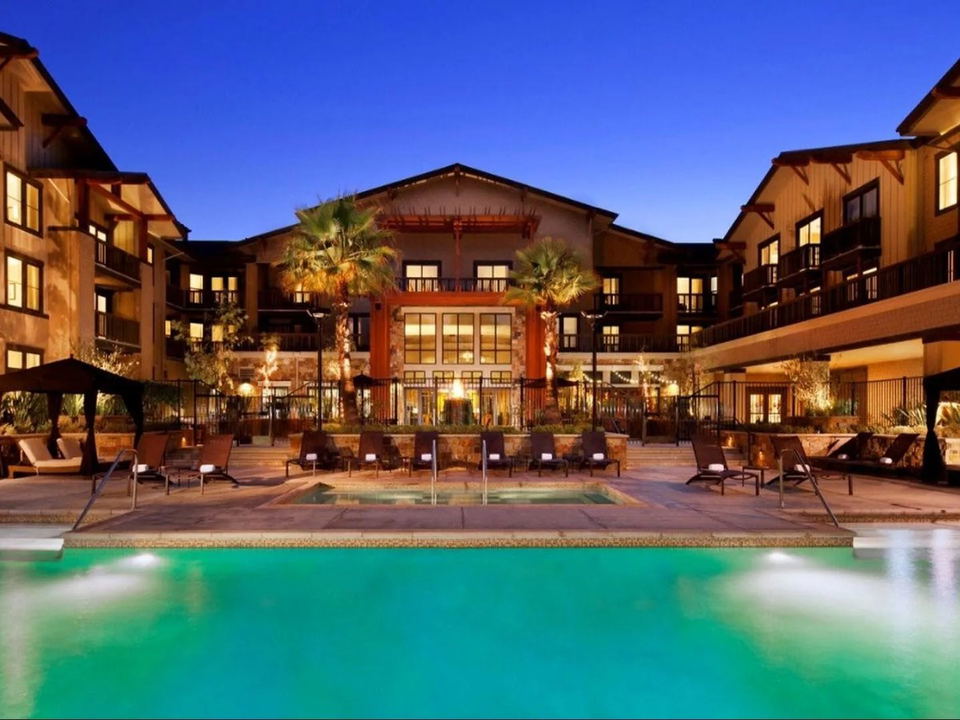 An evening exterior view of the Westin Verasa Napa, featuring a large illuminated swimming pool, lounge chairs, palm trees, and warm-lit buildings.