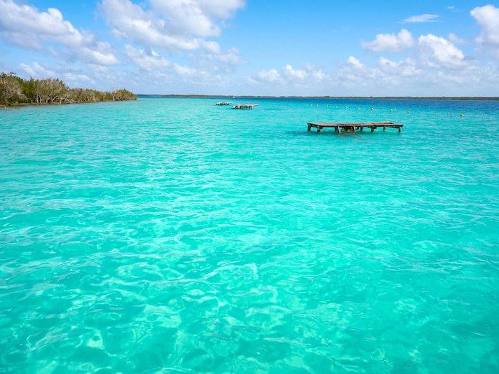 Wide landscape view of turquoise lagoon water with a wooden dock extending into the frame and distant shoreline with vegetation, published on a Bacala