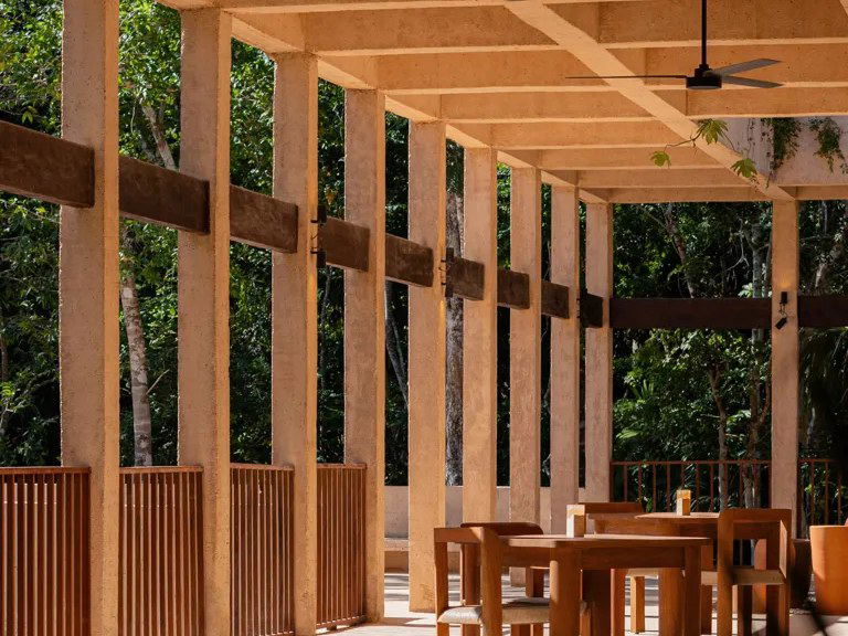 Open-air dining pavilion with wooden pergola structure, ceiling fans, and wooden furniture set against jungle foliage