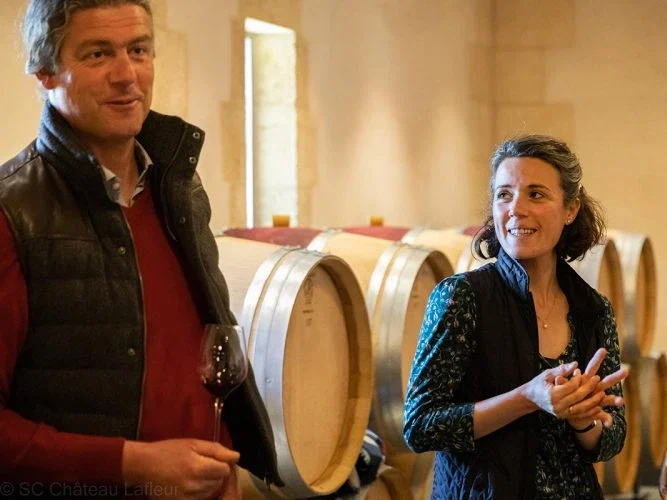 Baptiste Guinaudeau holding a glass of red wine and Julie Guinaudeau smiling with hands clasped, standing among oak barrels in the Château Lafleur cellar.