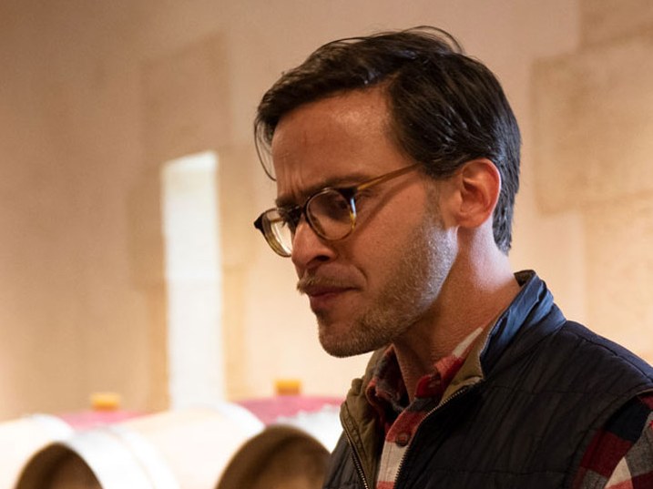 A man with dark hair and glasses wearing a dark jacket over a red plaid shirt studies rolled blueprints or plans on a table.