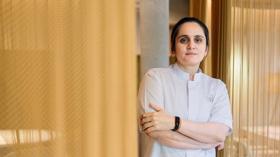 A woman wearing a white chef's coat with an embroidered logo stands with arms crossed in a modern interior space with warm-toned curtains.