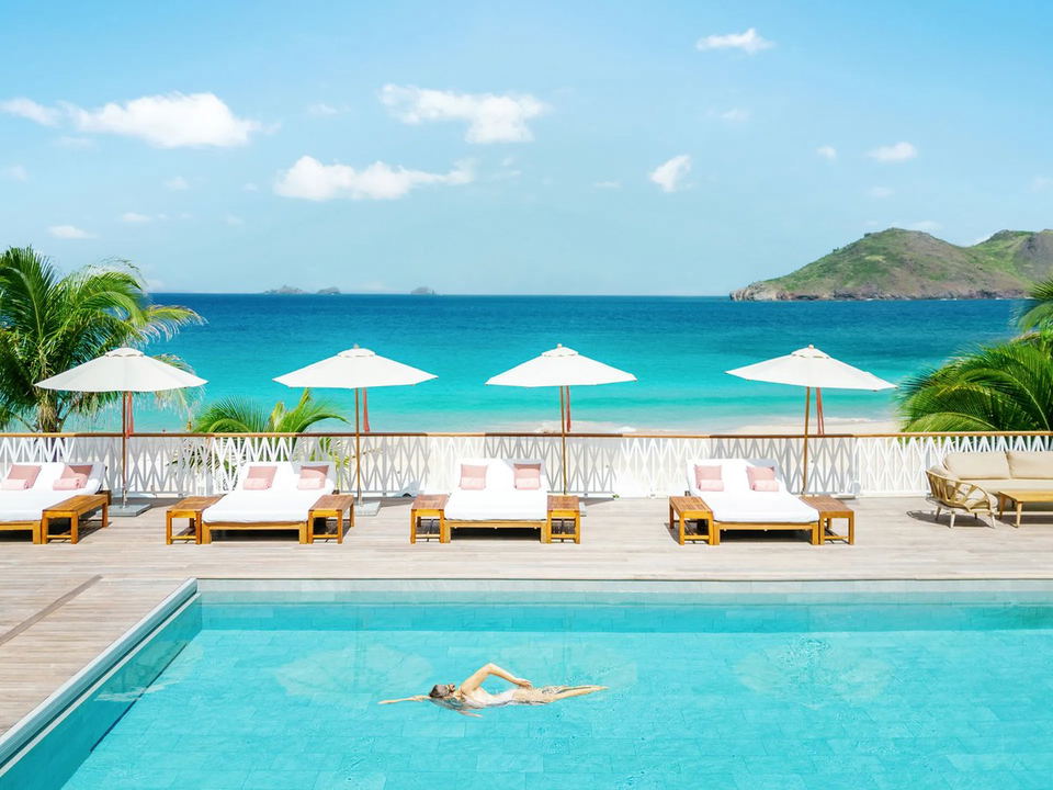 A luxury beachfront pool deck with lounge chairs, white umbrellas, palm trees, and turquoise ocean with an island in the distance; the source page (Co