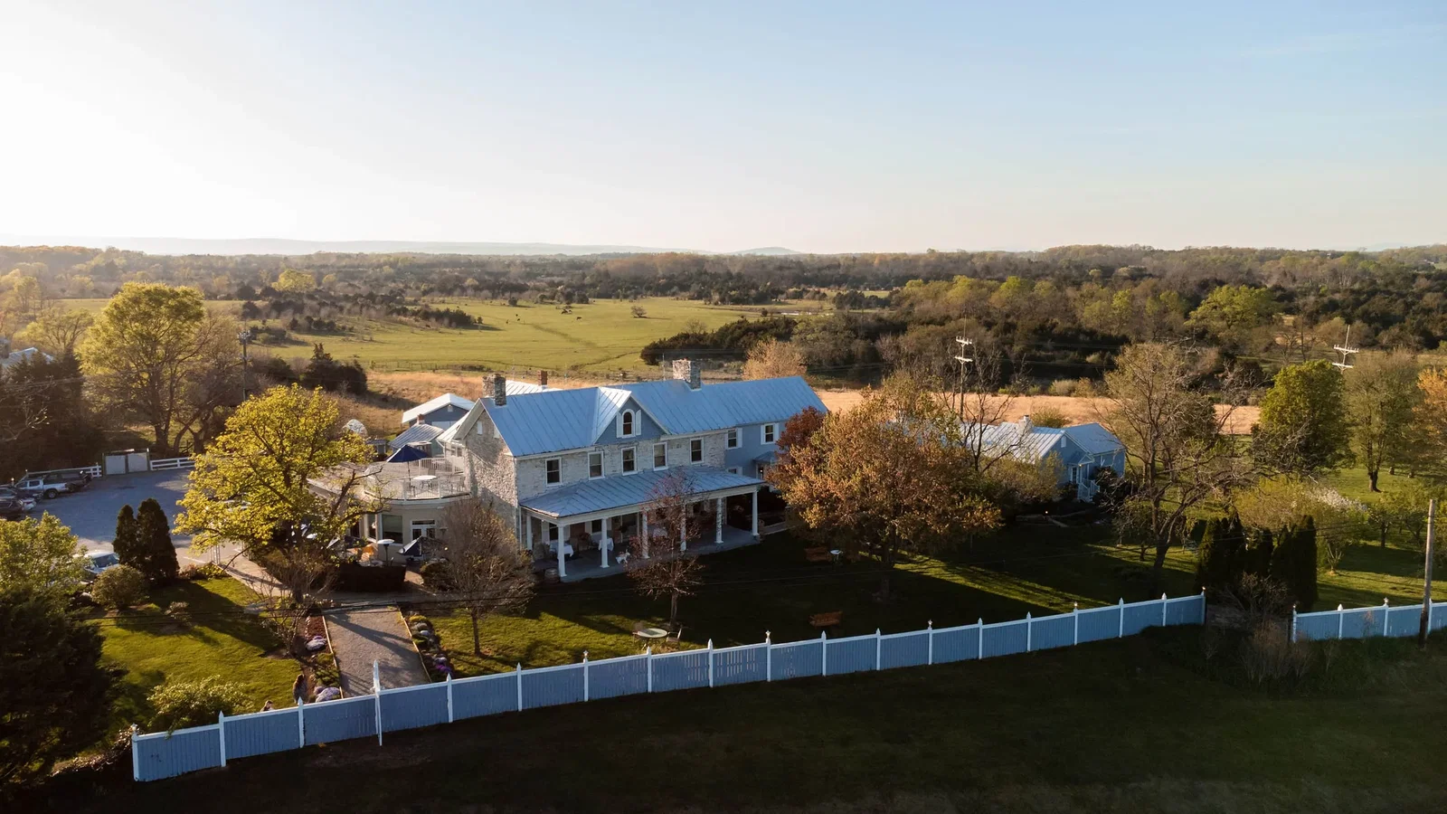 White farmhouse with blue metal roof surrounded by white picket fencing on expansive rural property with rolling pastures and tree-lined horizons.