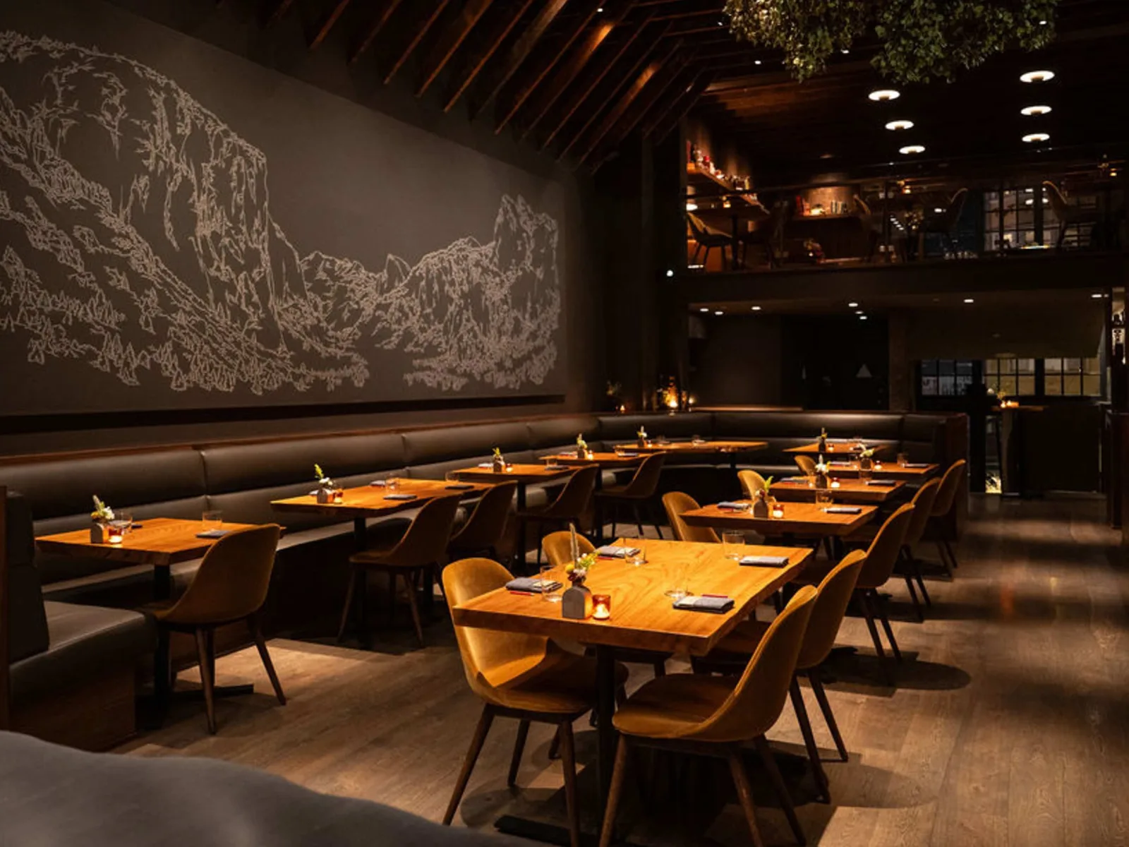 A modern restaurant dining room with wooden tables, golden chairs, dark leather seating along the wall, and a large illuminated landscape artwork on the back wall.