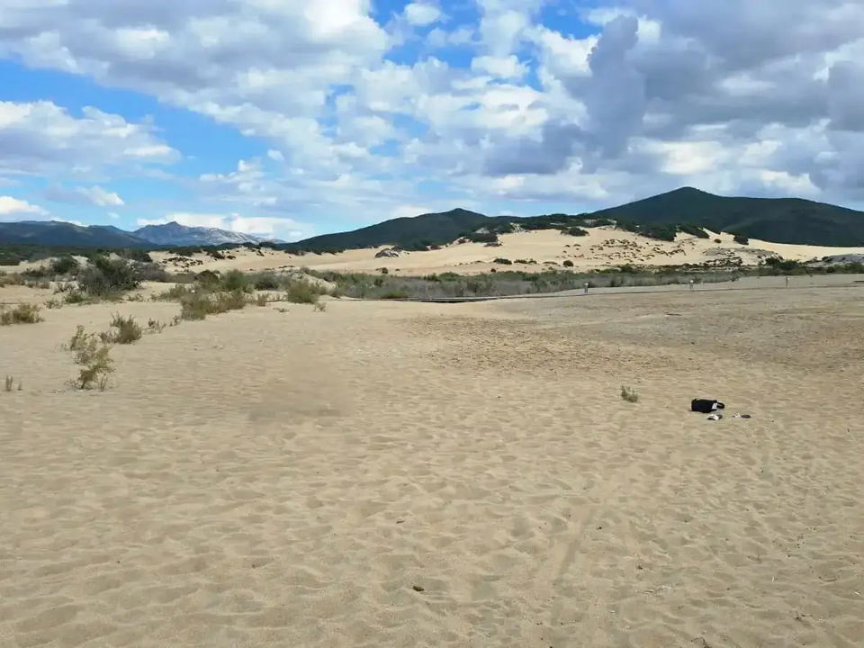 A sandy dune landscape with sparse vegetation under a partly cloudy sky, characteristic of the Piscinas region.