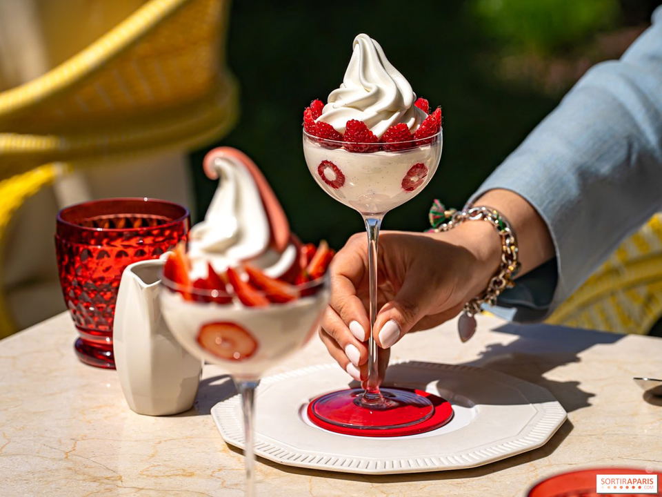 A person's hand holds a stemmed glass filled with soft-serve ice cream, raspberries, and cream, with another similar dessert out of focus in the
