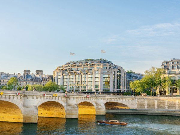 An elevated riverside view of the Cheval Blanc Paris building, formerly La Samaritaine, overlooking the Seine with a stone bridge in the foreground.