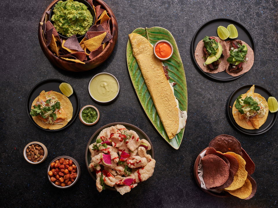 A flat-lay overhead composition of Mexican-style dishes including tacos, guacamole with chips, chicharrones, and various sauces arranged on dark plate
