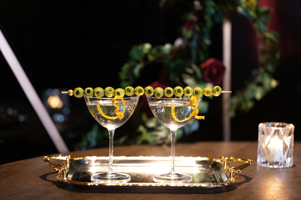 Two elegant coupe cocktails garnished with olives and lemon twists on a gold tray, photographed in a restaurant setting.